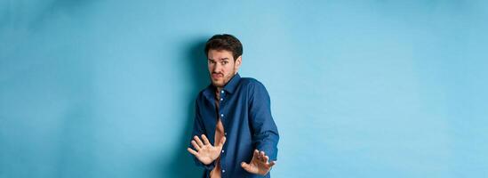 Timid young man asking to keep away, stretch out hands defensive and step back, refusing something disgusting, standing on blue background photo