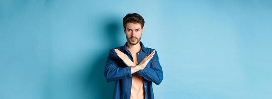 Serious caucasian man tell no, showing cross stop gesture and looking confident at camera, prohibit something bad, standing on blue background photo