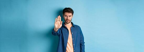 Upset man frowning and asking to stop, stretch out hand to prohibit or disagree with something bad, standing on blue background photo