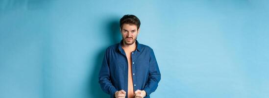 Angry and pissed-off man clench fists and stare with disdain at camera, trying to keep himself together, look with rage, standing on blue background photo