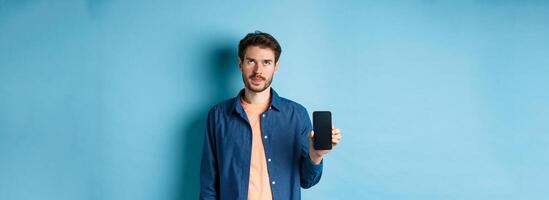 Annoyed young guy showing empty smartphone screen and roll eyes up bothered, standing on blue background photo