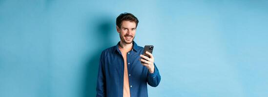 Attractive caucasian man using mobile phone, looking happy and smiling at camera, standing on blue background photo