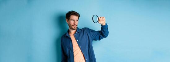 Image of young man searching for something, looking at empty space with magnifying glass, standing on blue background photo