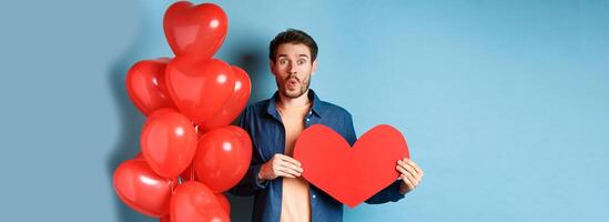 Valentines day concept. Boyfriend in love looking amazed at camera, holding red paper heart and standing with romantic balloon, blue background photo
