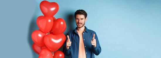 Happy valentines day. Confident man pointing fingers at camera and smiling, standing with balloons on blue background photo