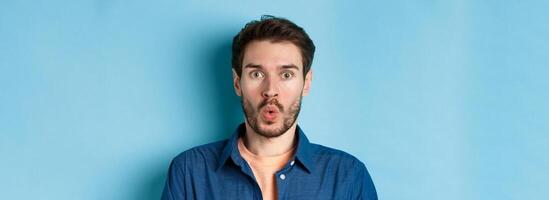 Close up portrait of amazed and surprised man saying wow, staring impressed at camera, checking out cool offer, standing on blue background photo