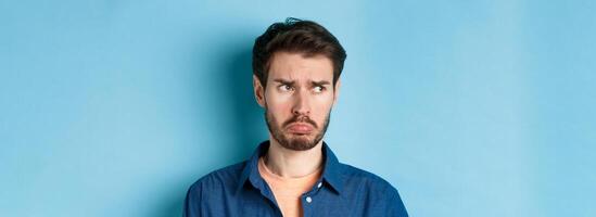 Miserable and disappointed guy acting childish, sulking and looking left at empty space with offended face, standing on blue background photo