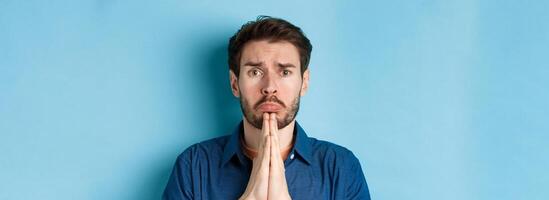 Sad young man begging for help, apoligizing and sobbing miserable, standing on blue background photo