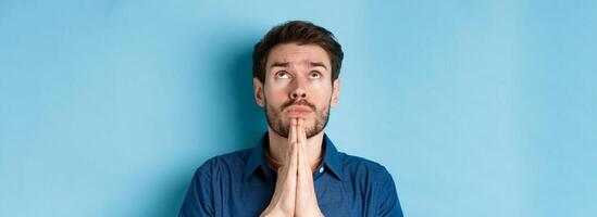 Close up shot of hopeful guy pleading God, holding hands in begging gesture and looking up in sky, standing on blue background supplicating photo