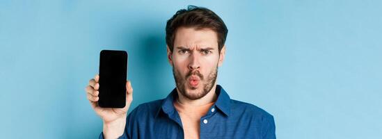 Shocked and confused young man frowning bothered, complaining at something on mobile phone, showing empty screen, standing on blue background photo