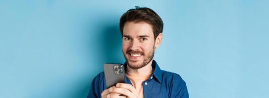 Close up of young guy with beard, smiling happy at camera and holding mobile phone, standing on blue background photo