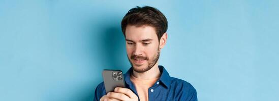 Close up portrait of happy young man reading mobile sreen, networking in smartphone, standing on blue background photo