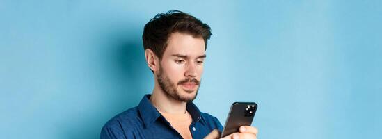 Close up of handsome bearded man reading mobile phone message, looking at screen serious, standing on blue background photo