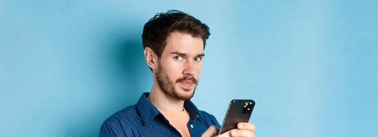 Close-up of handsome european guy using smartphone and looking at camera, standing on blue background photo