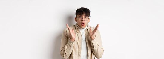 Portrait of surprised hipster guy in glasses checking out awesome promo, say wow and gasping amazed, raising hands up with disbelief, standing on white background photo