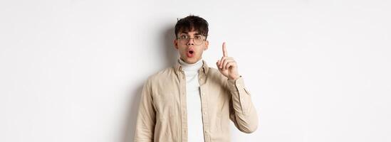 Portrait of young man student pitching an idea, raising finger up and say suggestion, have a plan, standing on white background in glasses and casual clothes photo