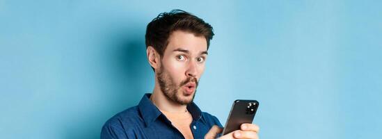 Close up of surprised caucasian man say wow, looking amused at camera, holding smartphone, standing on blue background photo