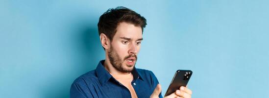 Close-up of man looking shocked and reading message on smartphone, gasping and stare startled at screen, standing on blue background photo