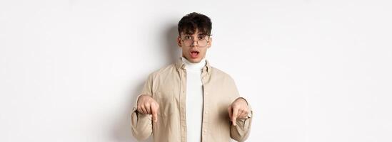 Shocked attractive young man showing something awesome, drop jaw and gasping in awe, pointing fingers down, standing on white background photo
