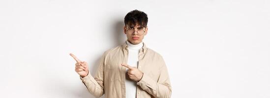 Angry young man frowning and pointing at upper right corner banner, complaining and looking disappointed, standing on white background photo