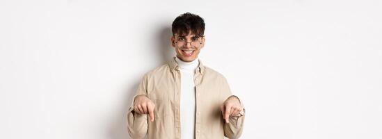 Stylish hipster male model in glasses pointing fingers down, smiling pleased at camera, recommending product, standing on white background photo