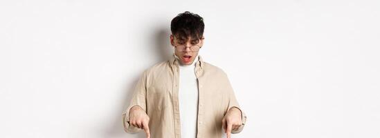Amazed young guy in glasses look and point down, open mouth and say wow impressed, checking out discount offer, standing on white background photo