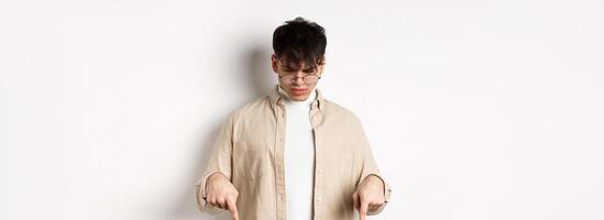 Confused frowning guy in glasses looking, pointing fingers down at something strange, standing puzzled on white background photo