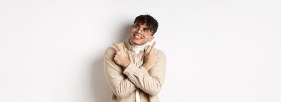 Portrait of handsome smiling guy in glasses pointing fingers sideways, made his choice and look at upper right corner logo, standing on white background photo
