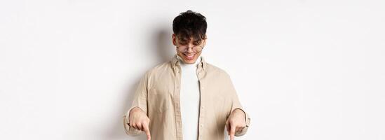 Stylish modern guy in glasses pointing, looking down with happy smile, showing banner, standing on white background photo