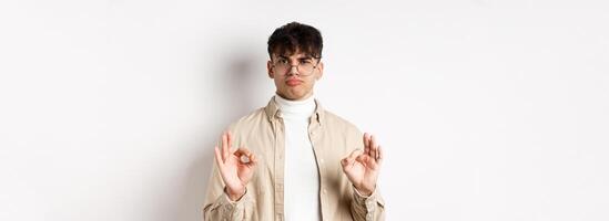 Not bad. Satisfied young man showing okay gestures and nod in approval, praise good work, pleased with results, standing on white background photo