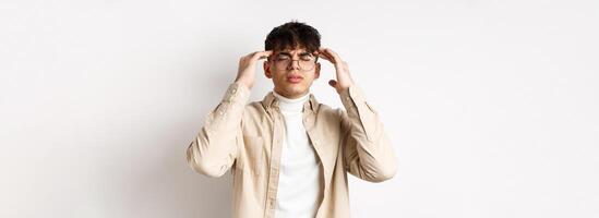 Image of young man in glasses having headache, touching head temples and frowning from painful migraine, feel dizzy, standing on white background photo