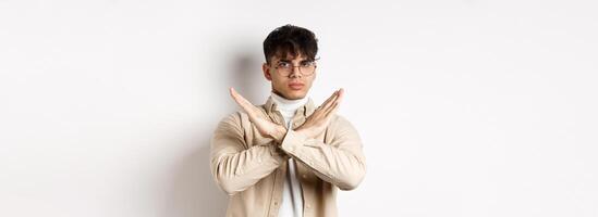 Image of serious young guy in glasses say no, showing cross gesture, make stop sign to disagree or prohibit something, standing on white background photo