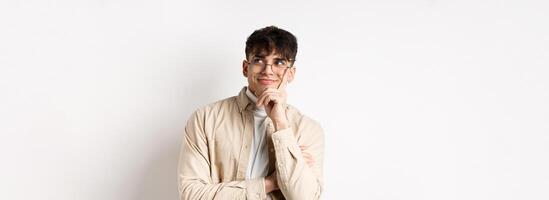 Image of happy dreamy young man imaging something good, smiling and looking at upper right corner thoughtful, standing on white background photo