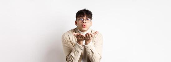 Cute boyfriend sending air kiss at camera on happy Valentines day, looking tender at lover, standing in casual clothes and glasses on white background photo