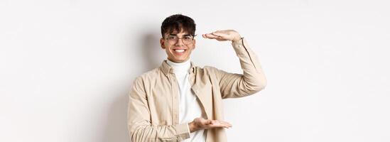 Real people. Happy modern guy in glasses showing big size, shaping box and smiling, showing something large, standing on white background photo