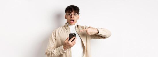Real people. Amazed young guy checking out online offer on mobile phone, pointing at smartphone screen with excited face, standing on white background photo