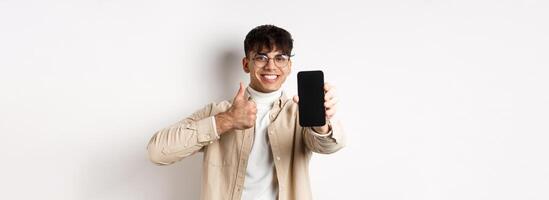 Portrait of handsome young man in glasses, showing empty smartphone screen and thumb up, recommending online app or store, standing pleased on white background photo