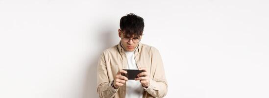 Handsome young man stare at mobile screen and playing game on smartphone, standing on white background photo