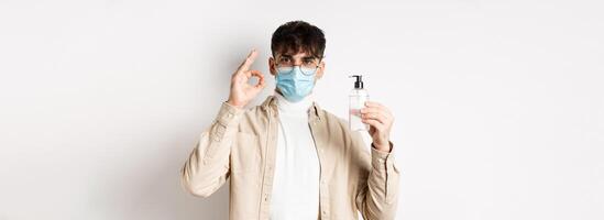 Health, covid and quarantine concept. Portrait of natural guy in glasses and face mask showing bottle of good hand sanitizer, make okay sign, recommending product, white background photo