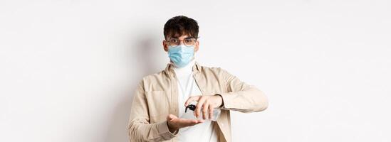 Health, covid and quarantine concept. Young hispanic guy in glasses and face mask using hand sanitizer, apply antiseptic and look at camera, standing on white background photo