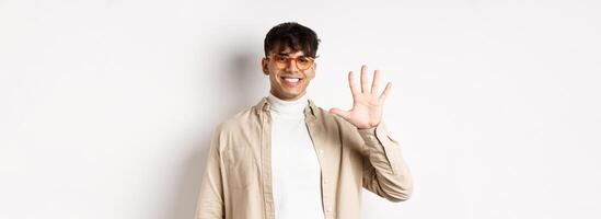 Modern guy in glasses and stylish outfit, showing five fingers number and smiling, making order, standing on white background photo