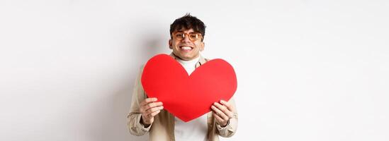 Cheerful young man smiling and looking thrilled at camera, showing big red heart cutout on Valentines day, making romantic gift to lover, white background photo