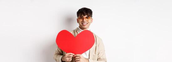 Handsome hipster guy with sunglasses and earring, waiting for true love on Valentines day, holding big red heart and smiling, standing over white background photo