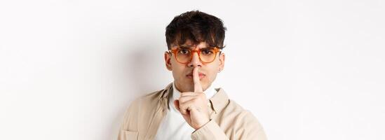 Close-up of grumpy young guy in glasses frowning and shushing, make taboo gesture with finger shuting lips, tell to be quiet, standing angry on white background photo