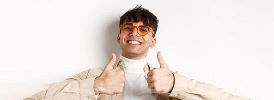 Close-up of satisfied happy guy showing thumbs up and smiling with white teeth, wearing eyewear, standing on white background photo