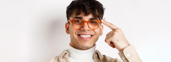 Headshot of smiling happy man showing new eyewear frame, pointing at glasses and looking satisfied, standing on white background photo