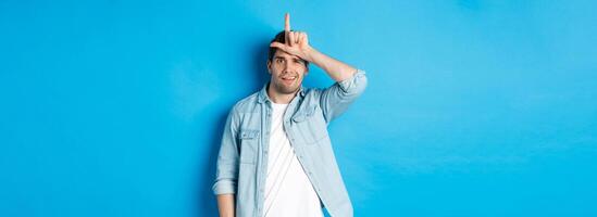 Rude guy mocking person for losing, showing loser sign on forehead and looking with dismay, standing against blue background photo