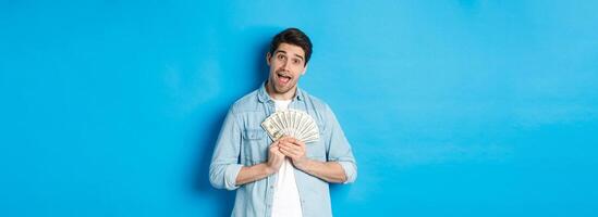 Smiling greedy guy hugging money and smiling, unwilling to share, standing over blue background photo