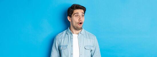 Close-up of surprised handsome man looking impressed left, open mouth fascinated, wearing casual outfit, standing over blue background photo