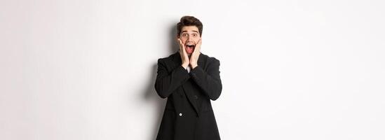 Portrait of surprised handsome businessman in suit, reacting to holidays promo, looking amazed at camera, standing over white background photo